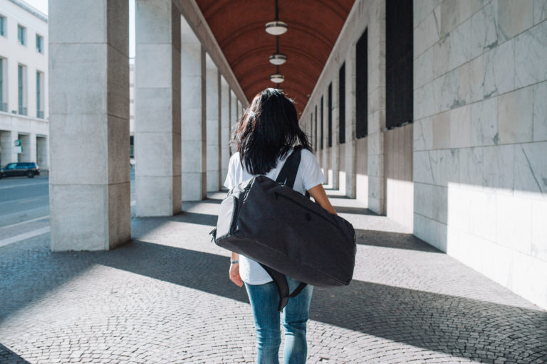 A woman carries a duffle bag in a portico.