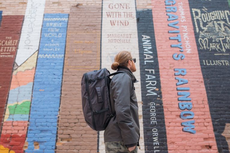Man wearing travel jacket and backpack near an interesting wall